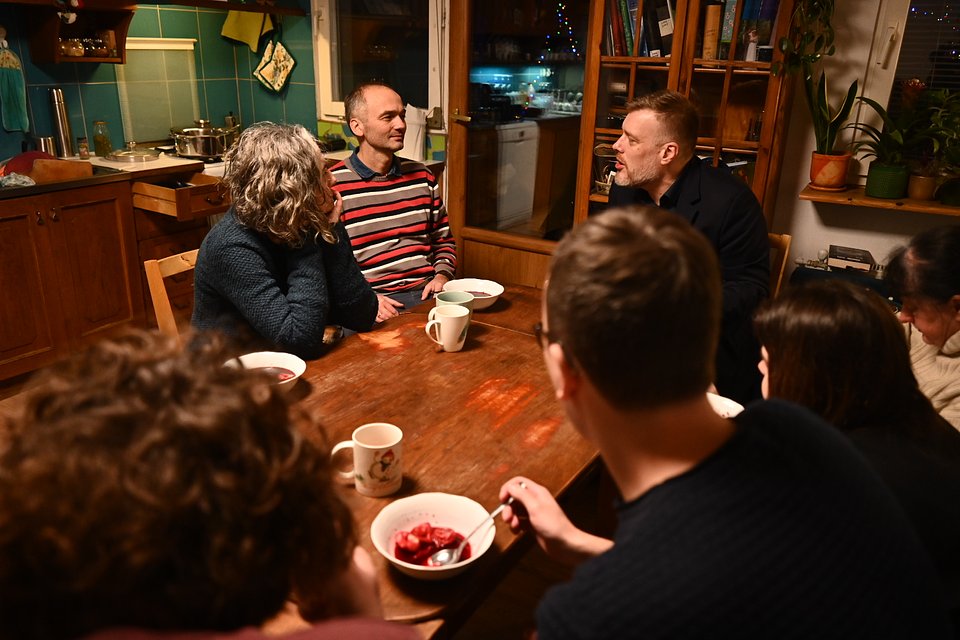 Na zdjęciu widzimy przytulne, domowe wnętrze – kuchnio-jadalnię z drewnianym stołem, otwartymi półkami, zielonymi płytkami na ścianie i regałami z książkami. Przy stole siedzi Adrian Zandberg, rozmawiający z grupą mieszkańców Osiedla Maltańskiego w Poznaniu.  Zandberg, ubrany w ciemny płaszcz, siedzi po prawej stronie stołu i prowadzi zaangażowaną rozmowę z osobami siedzącymi naprzeciwko. Jeden z rozmówców, mężczyzna w pasiastym swetrze, odpowiada z lekkim uśmiechem. Inni uczestnicy spotkania siedzą wokół stołu, niektórzy z pochylonymi głowami, wsłuchując się w rozmowę. Na stole stoją kubki i miseczki z jedzeniem, co podkreśla nieformalną, serdeczną atmosferę domowego spotkania.  Zdjęcie oddaje kameralny charakter rozmowy z mieszkańcami – pełnej uwagi, otwartości i troski o codzienne sprawy społeczności lokalnej.