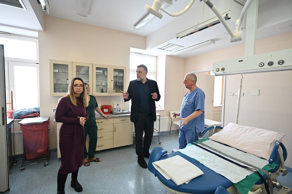 Na zdjęciu znajduje się wnętrze szpitalnej sali – jasne pomieszczenie z medycznym wyposażeniem, w tym specjalistycznym łóżkiem porodowym z pasami i aparaturą nad nim. W sali obecne są cztery osoby:  – Po lewej stronie stoi Marcelina Zawisza, ubrana w bordową sukienkę z długim rękawem i czarne buty. Ma długie, jasne włosy i okulary, patrzy w stronę fotografa z poważnym wyrazem twarzy. – Za nią stoi kobieta w zielonym uniformie medycznym – najprawdopodobniej członkini personelu pielęgniarskiego. – W centrum kadru znajduje się Adrian Zandberg, w czarnej marynarce i ciemnym ubraniu, gestykulujący i rozmawiający z mężczyzną w niebieskim stroju medycznym. – Po prawej stronie, przy łóżku, stoi lekarz w szpitalnym uniformie – to prawdopodobnie jeden z pracowników placówki.  Zdjęcie ukazuje wizytę polityków partii Razem w szpitalu w Płońsku, prawdopodobnie w ramach działań kontrolnych, interwencji poselskiej lub kampanii na rzecz ochrony zdrowia. Atmosfera jest rzeczowa i profesjonalna, a gesty wskazują na prowadzenie rozmowy dotyczącej warunków pracy lub potrzeb placówki.