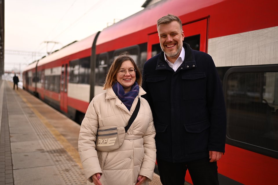 Na zdjęciu widzimy Paulinę Matysiak i Adriana Zandberga stojących razem na peronie dworca PKP w Kutnie. W tle znajduje się czerwono-biały pociąg, który właśnie zatrzymał się przy peronie. Oboje są uśmiechnięci, sprawiają wrażenie zadowolonych i gotowych do podróży lub właśnie z niej wrócili.  Paulina Matysiak ma na sobie jasną, pikowaną kurtkę i nosi charakterystyczną saszetkę z logiem PKP przewieszoną przez ramię. Na szyi ma fioletowy szalik, a na twarzy okulary i uśmiech. Adrian Zandberg ubrany jest w ciemną kurtkę i białą koszulę, również promienieje uśmiechem.  Zdjęcie zostało wykonane przez Dawida Majewskiego z Razem i przeznaczone jest do publicznego wykorzystania. Kompozycja ujęcia podkreśla zaangażowanie polityków w sprawy transportu publicznego i bliskość z codziennością mieszkańców – dworzec, pociągi, wspólne działania.