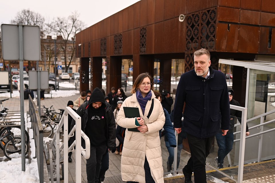 Na zdjęciu widzimy Paulinę Matysiak i Adriana Zandberga, którzy w towarzystwie grupy osób wychodzą po schodach spod Dworca PKP w Kutnie. W tle widoczna jest charakterystyczna, nowoczesna elewacja budynku z ażurowymi panelami i zimowy krajobraz – śnieg zalegający na ziemi oraz ludzie ubrani w ciepłe kurtki.  Paulina Matysiak idzie na czele, trzyma zieloną teczkę i nosi jasny płaszcz oraz fioletowy szalik. Obok niej Adrian Zandberg, ubrany w ciemną kurtkę, rozmawia z nią podczas wspólnego wejścia po schodach. Za nimi podąża kilkanaście osób, w tym osoby z przypinkami partii Razem.  Zdjęcie autorstwa Dawida Majewskiego z Razem, przeznaczone do wykorzystania, ukazuje polityków blisko ludzi i codziennych miejsc, takich jak infrastruktura transportowa, podkreślając ich zaangażowanie w sprawy publiczne i lokalne społeczności.