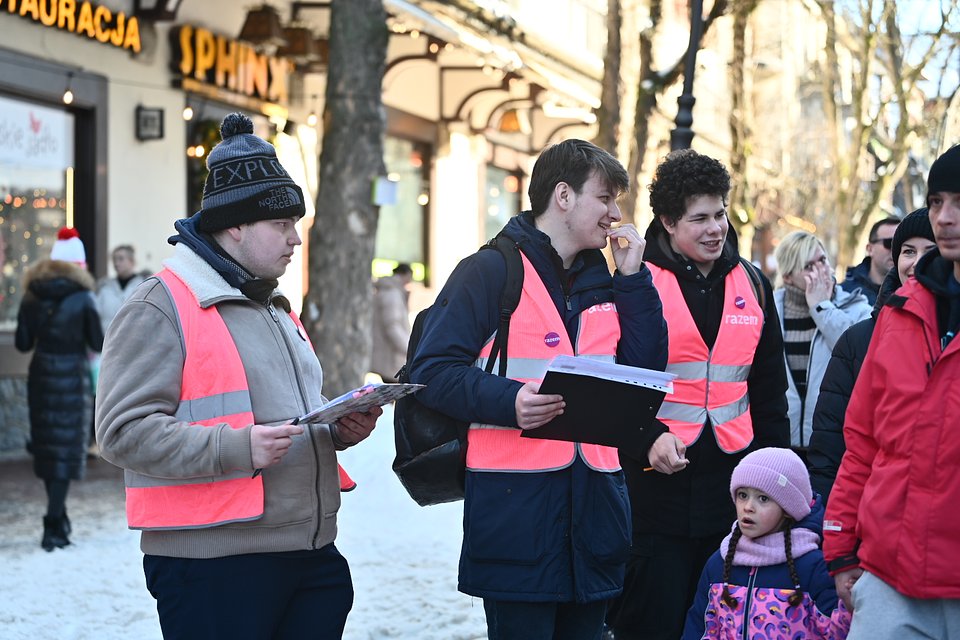 Członkowie młodzieżówki Razem podczas zbiórki podpisów w Zakopanem. Fot. Dawid Majewski z Razem Na zdjęciu widzimy grupę młodych osób – członków młodzieżówki partii Razem – prowadzących zbiórkę podpisów na ulicy w Zakopanem. Jest zima, na chodnikach leży śnieg, a wszyscy są ubrani w ciepłe ubrania. Trójka młodych mężczyzn nosi charakterystyczne odblaskowe kamizelki w kolorze różowym z napisem „razem”. Każdy z nich trzyma segregator lub kartkę – prawdopodobnie z listami do podpisu. Jeden z nich, w czapce z napisem „EXPLORE”, zwraca się do kogoś poza kadrem. Drugi – w granatowej kurtce – uśmiecha się i ma długopis w dłoni, gotowy do zbierania podpisów. Trzeci, z kręconymi włosami, również się uśmiecha, tworząc pogodną atmosferę. Obok nich stoi mężczyzna w czerwonej kurtce oraz mała dziewczynka w różowo-fioletowej kurtce i czapce z pomponem – trzyma kogoś za rękę i patrzy z zaciekawieniem na zbierających. W tle widać szyld restauracji „Sphinx” i zimową atmosferę Krupówek – głównego deptaka Zakopanego. Zdjęcie oddaje zaangażowanie młodych ludzi w kampanię polityczną oraz życzliwy kontakt z mieszkańcami i turystami.