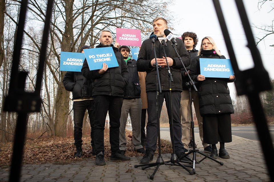 Na zdjęciu widzimy Adriana Zandberga podczas konferencji prasowej w miejscowości Świerk. Stoi na chodniku przy lesie, przemawiając do kilku mikrofonów ustawionych przed nim (widoczne są m.in. mikrofony TVN24 i Polsat News). Ubrany jest w czarną zimową kurtkę i mówi z zaangażowaniem, gestykulując dłońmi.  Za nim stoi grupa osób wspierających, trzymających niebieskie i różowe tablice z hasłami kampanii: „Adrian Zandberg” i „Po twojej stronie”. W tle widoczne są bezlistne drzewa, sugerujące, że zdjęcie wykonano zimą lub późną jesienią. Asfaltowa droga i leśne otoczenie tworzą kontekst przestrzeni publicznej poza centrum miasta – być może nawiązując do spraw lokalnych, infrastruktury, transportu lub ochrony środowiska.  Całość ujęcia wskazuje na formalne wystąpienie polityczne, a obecność kamery (jej statywy widoczne na pierwszym planie) podkreśla medialny charakter wydarzenia.