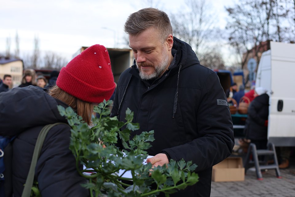 Na zdjęciu widzimy Adriana Zandberga podczas spotkania z mieszkańcami na lokalnym targowisku. Ubrany jest w czarną zimową kurtkę z kapturem, stoi przy stoisku lub wśród kupujących, z lekko pochyloną głową – prawdopodobnie rozmawia lub zbiera podpisy.  Na pierwszym planie znajduje się kobieta w czerwonej czapce i ciemnym płaszczu z torbą na ramieniu, zasłonięta częściowo przez świeżą natkę pietruszki, co nadaje zdjęciu naturalnego i codziennego charakteru. W tle widać innych ludzi, samochody dostawcze i pudełka, typowe dla miejskiego targu.  Atmosfera zdjęcia jest swobodna i bezpośrednia – Zandberg nawiązuje kontakt z mieszkańcami w ich codziennym otoczeniu, co podkreśla jego dostępność i zaangażowanie w sprawy lokalne.