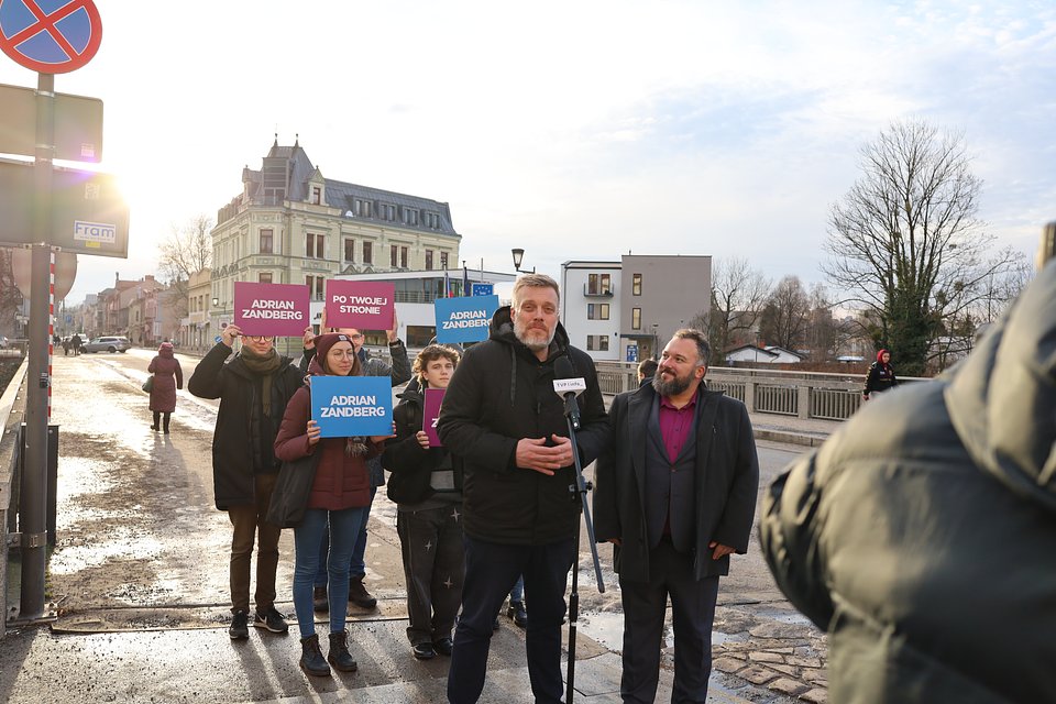 Adrian Zandberg na Moście Przyjaźni w Cieszynie, po prawej stronie radny Cieszyna Paweł Jasiński Na zdjęciu widzimy Adriana Zandberga, który przemawia podczas konferencji prasowej na tle zimowego krajobrazu, stojąc na Moście Przyjaźni w Cieszynie – symbolicznym przejściu granicznym łączącym Polskę i Czechy. Zandberg ubrany jest w ciemną kurtkę i mówi do mikrofonu TVP Gorzów, a obok niego stoi Paweł Jasiński, radny Cieszyna, ubrany w ciemny garnitur i koszulę w kolorze fuksji. Spogląda z uśmiechem w stronę Zandberga. Za nimi stoi grupa osób trzymających kolorowe tablice kampanijne: „Adrian Zandberg” i „Po twojej stronie” – w barwach niebieskich i fioletowych. W tle widać secesyjne kamienice Cieszyna oraz zimowe, rozświetlone słońcem niebo. Po lewej stronie kadru widać znak drogowy i przejście graniczne z charakterystycznym oznaczeniem. Atmosfera zdjęcia jest oficjalna, ale pogodna – pokazuje lokalne zaangażowanie kampanii wyborczej i obecność polityka w przestrzeniach symbolicznie ważnych dla współpracy i sąsiedztwa.
