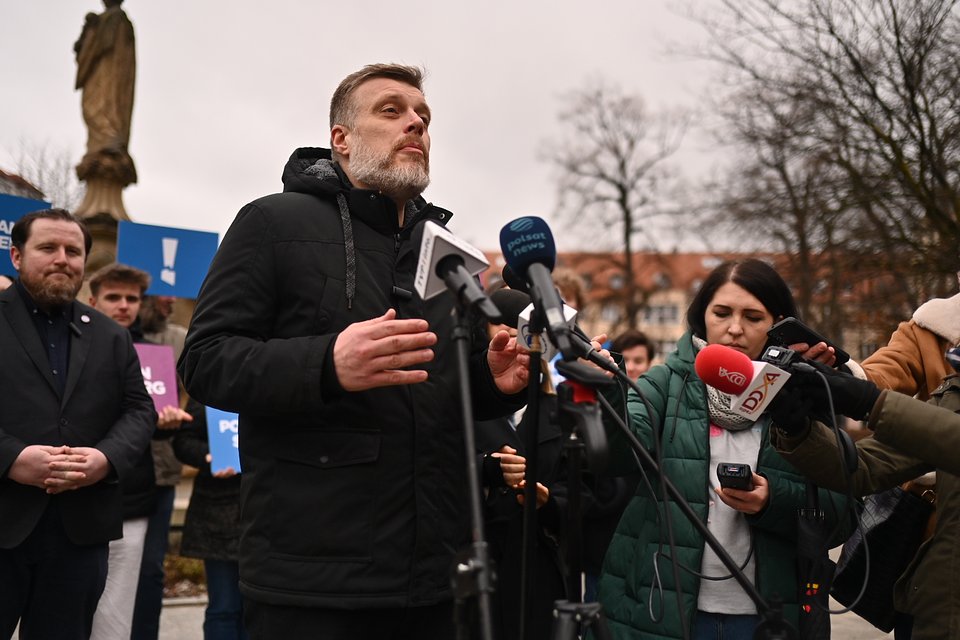 Adrian Zandberg podczas konferencji w Opolu. Fot. Dawid Majewski z Razem Na zdjęciu znajduje się scena z zewnętrznej konferencji prasowej, która odbywa się w pochmurny dzień. Na pierwszym planie stoi mężczyzna – to Adrian Zandberg – przemawiający do mikrofonów różnych mediów (widoczne są m.in. mikrofony Polsat News, TVP Info, Radio Zet). Ma krótko przystrzyżoną brodę, jasne włosy, a ubrany jest w czarną kurtkę z kapturem. Wydaje się być skupiony i zaangażowany w to, co mówi, gestykuluje dłońmi. Obok niego stoi kobieta z ciemnymi włosami, ubrana w zieloną kurtkę – trzyma telefon i mikrofon, najpewniej jest dziennikarką. W tle widać grupę ludzi – część z nich trzyma tablice z logotypem partii Razem i znakami wykrzyknika. Wśród nich są młodzi mężczyźni oraz inny polityk z brodą, w garniturze. Za zgromadzeniem widać nagie drzewa i klasyczne budynki z czerwonymi dachami, a także kamienną rzeźbę – najpewniej postać świętego – co sugeruje, że wydarzenie odbywa się na placu lub w parku w centrum miasta. Atmosfera jest poważna, a wydarzenie sprawia wrażenie politycznego wiecu lub oświadczenia dla mediów.