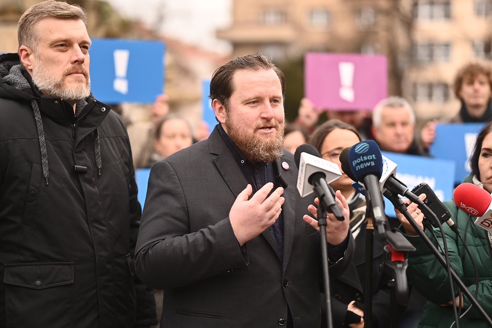 Jerzy Przystajko podczas konferencji prasowej z Adrianem Zandbergiem w Opolu. Fot. Dawid Majewski z Razem Na zdjęciu uchwycono moment podczas konferencji prasowej lub wiecu politycznego. W centrum kadru stoi Jerzy Przystajko, przemawiający do zgromadzonych dziennikarzy i mikrofonów (widoczne są m.in. mikrofony Polsat News i Radia Zet). Jest to mężczyzna z ciemnymi włosami i brodą, ubrany w ciemną marynarkę i ciemną koszulę. Wypowiada się z zaangażowaniem – trzyma ręce na wysokości klatki piersiowej w geście podkreślającym wypowiedź. Po lewej stronie, nieco w tle, stoi Adrian Zandberg w czarnej kurtce z kapturem, z charakterystyczną siwą brodą i poważnym wyrazem twarzy. Obaj politycy znajdują się na pierwszym planie i są wyraźnie głównymi postaciami wydarzenia. W tle widoczni są uczestnicy wydarzenia, trzymający tablice z dużymi wykrzyknikami na kolorowym tle – niebieskim i fioletowym – co jest elementem identyfikacji wizualnej partii Razem. Zgromadzeni ludzie są skupieni, uważnie słuchają przemówienia. Za nimi widać zabudowę miejską i drzewa, sugerujące, że wydarzenie ma miejsce na otwartej przestrzeni w centrum miasta. Atmosfera zdjęcia jest poważna i