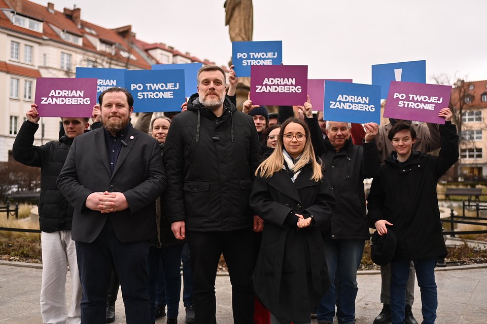 Od lewej Jerzy Przystajko, Adrian Zandberg, oraz Marcelina Zawisza podczas konferencji prasowej w Opolu. Fot. Dawid Majewski z Razem Na zdjęciu widać grupowe ujęcie uczestników wydarzenia politycznego – najprawdopodobniej wiecu poparcia lub ogłoszenia kampanii. W centrum kadru stoją trzy osoby: Jerzy Przystajko – po lewej stronie, w ciemnej marynarce, z brodą i delikatnym uśmiechem, trzyma ręce splecione przed sobą. Adrian Zandberg – w środku, w czarnej kurtce z kapturem, z poważnym wyrazem twarzy i charakterystyczną siwiejącą brodą. Marcelina Zawisza – po prawej stronie Zandberga, w czarnym płaszczu, z długimi jasnymi włosami i okularami. Również stoi spokojnie, patrząc wprost w obiektyw. Za nimi zgromadziła się grupa sympatyków, którzy trzymają tablice w barwach partii Razem – niebieskie i fioletowe, z hasłami takimi jak „Adrian Zandberg” oraz „Po twojej stronie”, a także z wykrzyknikiem – symbolem ugrupowania. Wszyscy są uśmiechnięci lub wyglądają na zaangażowanych, patrzą w stronę fotografa. Tłem są miejskie kamienice z czerwonymi dachami i rzeźba postaci świętego, co sugeruje, że zdjęcie zrobiono na placu w historycznej części miasta. Ujęcie ma charakter promocyjny i wspólnotowy – prezentuje solidarność i jedność kandydatów i ich zespołu.