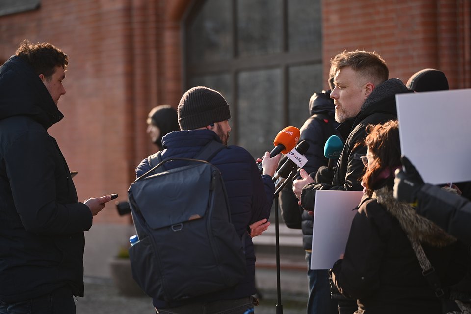 Na zdjęciu widzimy Adriana Zandberga podczas konferencji prasowej w Słupsku. Zandberg stoi zwrócony w stronę mikrofonów kilku redakcji – widoczne są m.in. pomarańczowy mikrofon Radia Zet, biały z logo TVP Info oraz turkusowy z innego medium. Ubrany jest w czarną kurtkę, a jego twarz jest oświetlona popołudniowym słońcem.  Przed nim zgromadzeni są dziennikarze – niektórzy trzymają smartfony lub notatniki, jeden z nich ma na sobie plecak i czapkę, inna osoba trzyma planszę lub baner. W tle widać ceglaną elewację budynku w stylu neogotyckim – prawdopodobnie ratusz lub inny gmach użyteczności publicznej.  Atmosfera jest poważna, ale spokojna – to typowy moment kontaktu polityka z mediami, podczas którego przekazuje ważne informacje lub komentuje bieżące sprawy. Zdjęcie podkreśla zaangażowanie Zandberga w działania terenowe i otwartość na rozmowę z prasą.