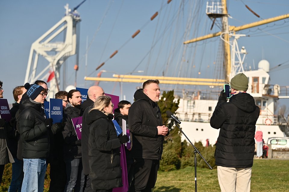 Na zdjęciu widzimy Adriana Zandberga, który przemawia podczas porannej konferencji prasowej w Gdyni. Stoi na trawniku, zwrócony w stronę kamery ustawionej na statywie – obsługiwanej przez osobę w zielonej czapce i czarnej kurtce.  Za Zandbergiem stoi grupa młodych ludzi, część z nich trzyma plakaty z hasłami kampanii: „Adrian Zandberg” oraz „Po Twojej stronie”. Wszyscy są ubrani w zimowe ubrania, a słońce jasno świeci, sugerując chłodny, ale pogodny dzień.  W tle widać cumujący żaglowiec szkolny „Dar Pomorza” oraz fragment portowego dźwigu. Obecność żaglowca i widok na morze jednoznacznie lokalizują wydarzenie na Nabrzeżu Pomorskim w Gdyni, popularnym miejscu konferencji i spotkań publicznych.