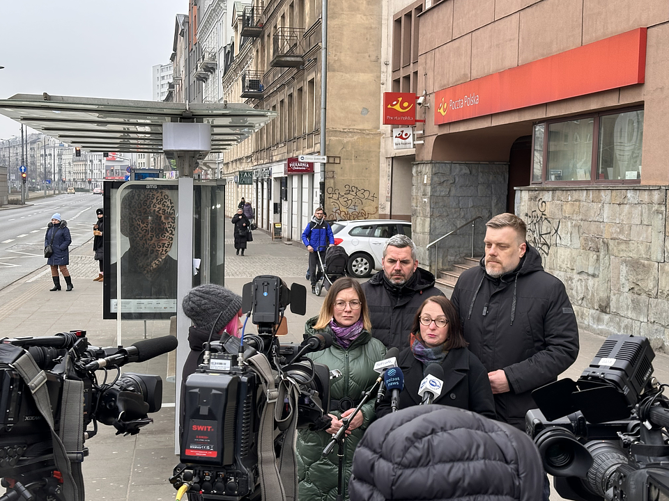 Na zdjęciu widzimy grupę polityków i działaczy partii Razem podczas konferencji prasowej przed oddziałem Poczty Polskiej. Stoją na chodniku przy ulicy w centrum miasta.  Na pierwszym planie, po lewej stronie kadru, znajduje się kilka kamer telewizyjnych i mikrofonów opatrzonych logotypami stacji takich jak TVN24, Polsat News, TVP Info. Przed kamerami stoją cztery osoby:  Paulina Matysiak (po lewej) ma blond włosy, okulary i zieloną, pikowaną kurtkę. Mówi do mikrofonów.  Maciej Konieczny (za nią) ma siwe włosy i czarną kurtkę.  Magdalena Madzia (z prawej strony Matysiak) ma ciemne włosy, okulary i ciemną kurtkę. Również przemawia.  Adrian Zandberg (najbardziej po prawej) ma jasne włosy i brodę, ubrany jest w czarną kurtkę, stoi z powagą.  W tle widoczny jest budynek Poczty Polskiej z czerwonym szyldem oraz stara kamienica z graffiti. Po lewej stronie zdjęcia stoi wiata przystanku autobusowego z plakatem w gablocie. Ulica jest pusta, a pogoda pochmurna i chłodna – ludzie mają na sobie zimowe ubrania.