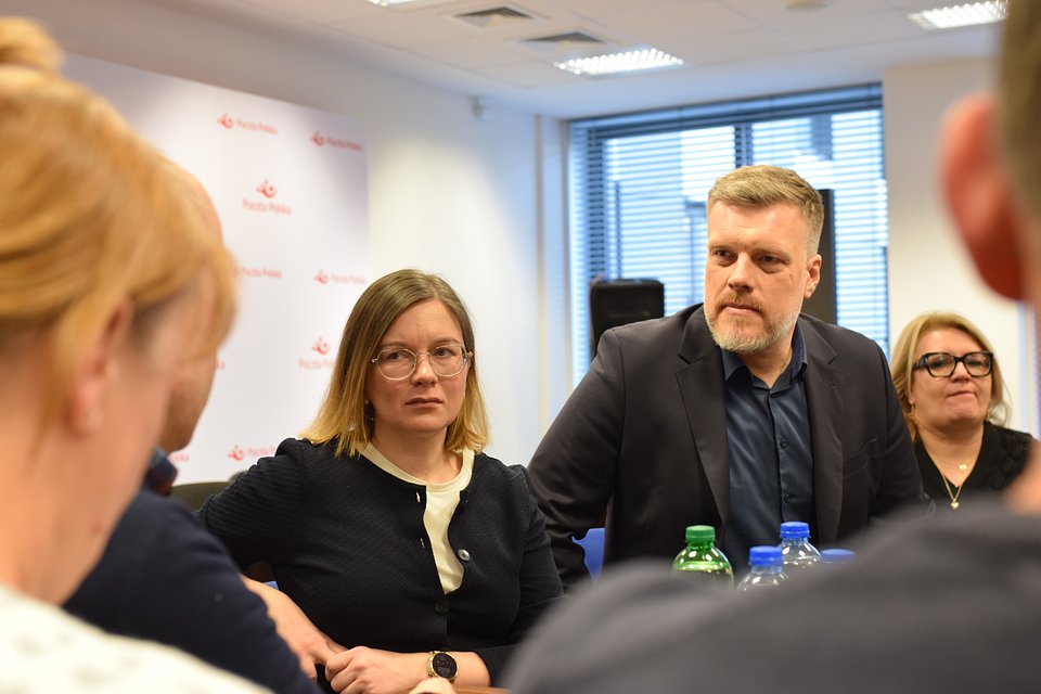 Na zdjęciu widzimy Paulinę Matysiak i Adriana Zandberga podczas rozmów z pracownikami Poczty Polskiej. Spotkanie odbywa się w siedzibie instytucji – w tle widoczne jest logo Poczty Polskiej na ściance konferencyjnej. Atmosfera spotkania jest poważna i skupiona, a rozmówcy siedzą przy stole z butelkami wody, co sugeruje dłuższą i merytoryczną dyskusję.  To zdjęcie świetnie oddaje zaangażowanie polityków partii Razem w sprawy pracownicze i dialog ze środowiskami zawodowymi.