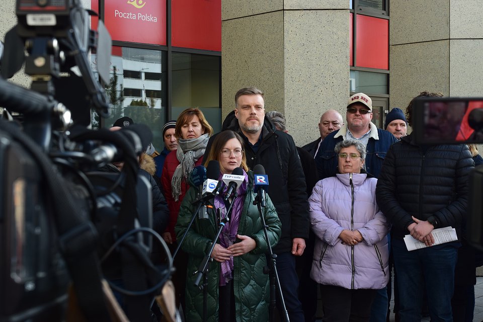 Na zdjęciu widzimy Paulinę Matysiak i Adriana Zandberga podczas konferencji prasowej zorganizowanej pod siedzibą Poczty Polskiej. Towarzyszy im grupa pracowników i działaczy związkowych, co podkreśla wagę poruszanych tematów – prawdopodobnie związanych z warunkami pracy, wynagrodzeniami lub przyszłością usług publicznych.  Kamera, mikrofony mediów i powaga wypowiedzi wskazują, że konferencja miała charakter interwencyjny i dotyczyła istotnych kwestii społecznych.