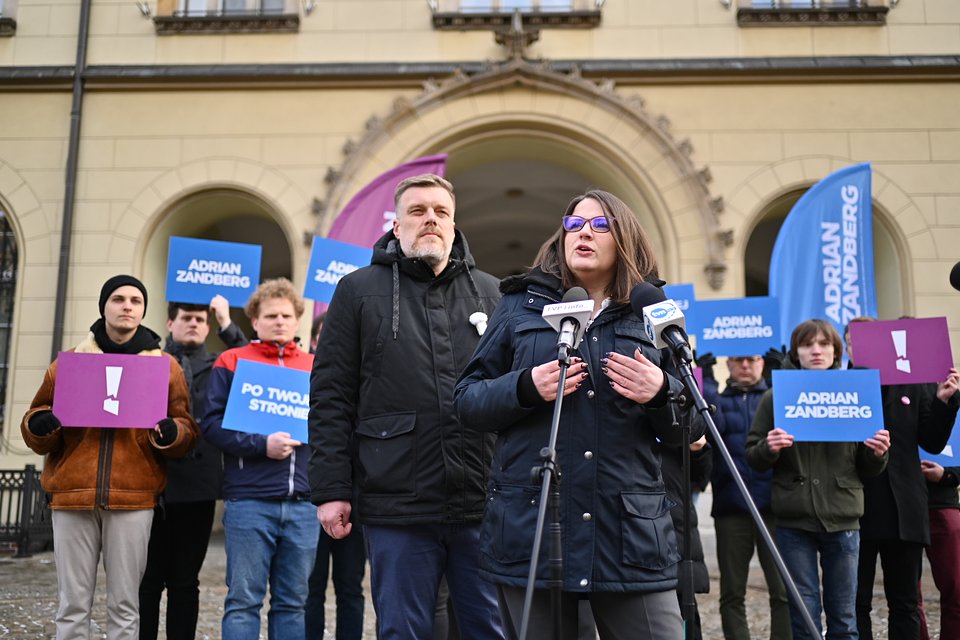Na zdjęciu widzimy Martę Stożek i Adriana Zandberga podczas konferencji prasowej we Wrocławiu. Marta Stożek przemawia do mikrofonów, stojąc na tle Adriana Zandberga i grupy wspierających ich osób trzymających plansze z hasłami kampanii: „Adrian Zandberg”, „Po twojej stronie” oraz charakterystyczne fioletowe plansze z wykrzyknikiem.  Tło stanowi zabytkowy budynek z arkadowym wejściem – prawdopodobnie siedziba instytucji publicznej. Otoczenie, stroje uczestników i bannery wskazują na wydarzenie o oficjalnym, kampanijnym charakterze.
