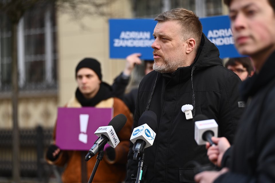 Na zdjęciu widzimy Adriana Zandberga podczas konferencji prasowej we Wrocławiu. Polityk przemawia na tle charakterystycznych niebieskich i fioletowych plansz z hasłami kampanii, m.in. „Adrian Zandberg” oraz „Po Twojej stronie”. Za nim stoją sympatycy i członkowie jego sztabu – część trzyma tablice z symbolami kampanii, w tym fioletowy wykrzyknik.  Zandberg ubrany jest w ciemną kurtkę z przypiętym mikrofonem reporterskim, co wskazuje na chłodną pogodę i formalny charakter wystąpienia. Na pierwszym planie widoczne są mikrofony kilku redakcji, w tym TVP Info i TVN24.