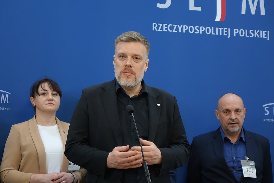 Na zdjęciu widzimy Adriana Zandberga podczas konferencji prasowej w Sejmie Rzeczypospolitej Polskiej. Tło z wyraźnym napisem „SEJM RZECZYPOSPOLITEJ POLSKIEJ” oraz logotypami instytucji wskazuje na oficjalny charakter wydarzenia.  Zandberg występuje na pierwszym planie, ubrany na czarno, co podkreśla powagę przekazu. Mikrofon umieszczony na mównicy sugeruje, że przemawia do mediów. Towarzyszą mu dwie osoby – kobieta w beżowej marynarce oraz mężczyzna w granatowej koszuli – oboje z identyfikatorami, co może świadczyć o tym, że są jego współpracownikami lub współuczestnikami wydarzenia.