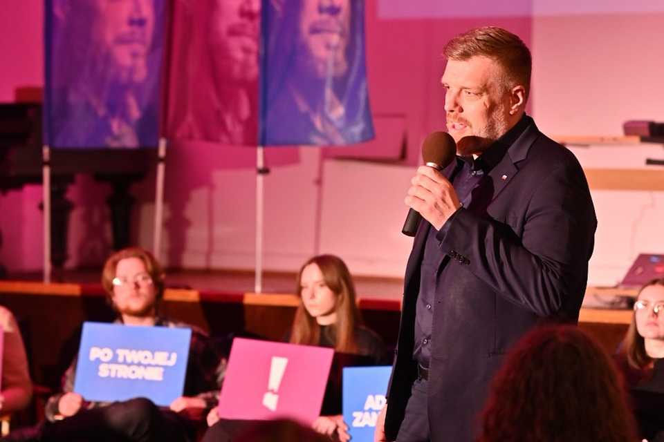 Na zdjęciu widzimy Adriana Zandberga podczas spotkania z wyborcami w Łodzi. Wydarzenie odbywa się we wnętrzu — w sali o różowym oświetleniu, z charakterystycznymi banerami z jego wizerunkiem oraz hasłami kampanii „Po twojej stronie” i „Adrian Zandberg”.  Zandberg przemawia do zgromadzonej publiczności, trzymając mikrofon. Na scenie towarzyszą mu młodzi ludzie z transparentami, co wskazuje na mocne zaangażowanie młodego elektoratu w jego kampanię. Atmosfera spotkania wydaje się być bardziej kameralna i skoncentrowana na dialogu.