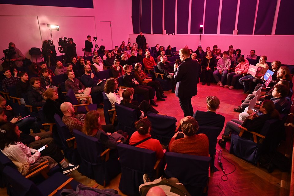 Na zdjęciu widzimy Adriana Zandberga przemawiającego podczas spotkania z wyborcami w Łodzi. Spotkanie odbywa się w zamkniętej sali o ciepłym, różowym oświetleniu. Publiczność otacza Zandberga w półkolu – to układ sprzyjający bezpośredniemu kontaktowi i rozmowie, który kojarzy się z otwartym, uczestniczącym stylem polityki.  Widać obecność kamer i mikrofonów, co sugeruje medialne zainteresowanie wydarzeniem. Z przodu siedzą uczestnicy z transparentami kampanii („Adrian Zandberg”, „Po twojej stronie”, wykrzyknik na niebieskim tle), a sam kandydat trzyma mikrofon i mówi z zaangażowaniem, co świadczy o dynamicznej interakcji z publicznością.