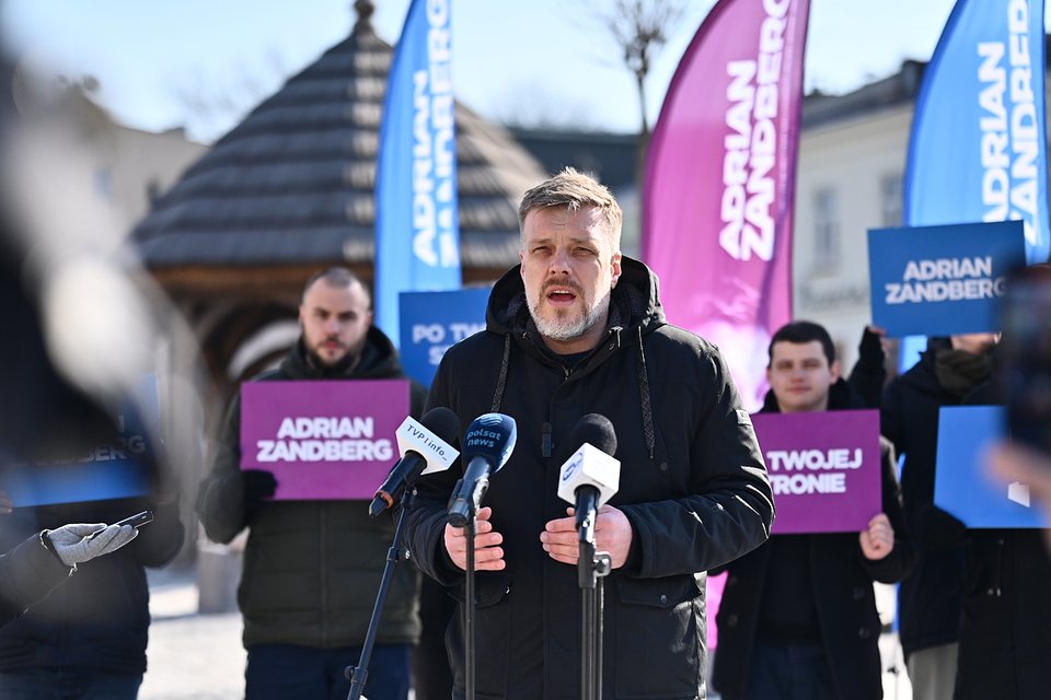 To zdjęcie przedstawia Adriana Zandberga podczas konferencji prasowej w Chełmie. Przemawia na tle fioletowo-niebieskich flag z jego nazwiskiem oraz haseł kampanii: „Po twojej stronie” i „Adrian Zandberg”. Obok niego widoczni są lokalni działacze i działaczki Razem, trzymający plansze wspierające kampanię.  W tle charakterystyczna altana z drewnianym dachem sugeruje, że wydarzenie miało miejsce na jednym z centralnych placów miasta — prawdopodobnie w obrębie rynku lub przy deptaku.