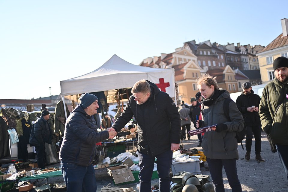To zdjęcie przedstawia Adriana Zandberga podczas spotkania z mieszkańcami Chełma, które odbyło się na lokalnym targowisku. Widać, że rozmowa z wyborcami to dla niego nie tylko obowiązek kampanijny, ale też okazja do bezpośredniego kontaktu i wymiany poglądów – uścisk dłoni i uśmiechy są wyrazem wzajemnego szacunku i otwartości.  W tle rozpoznawalne kramy z militariami oraz namiot z symbolem Czerwonego Krzyża tworzą ciekawy kontekst – być może odbywał się tam jarmark historyczny lub giełda kolekcjonerska. Miasto zyskało przez to wyjątkowy, lokalny klimat, który podkreślają również malownicze kamienice Chełma.