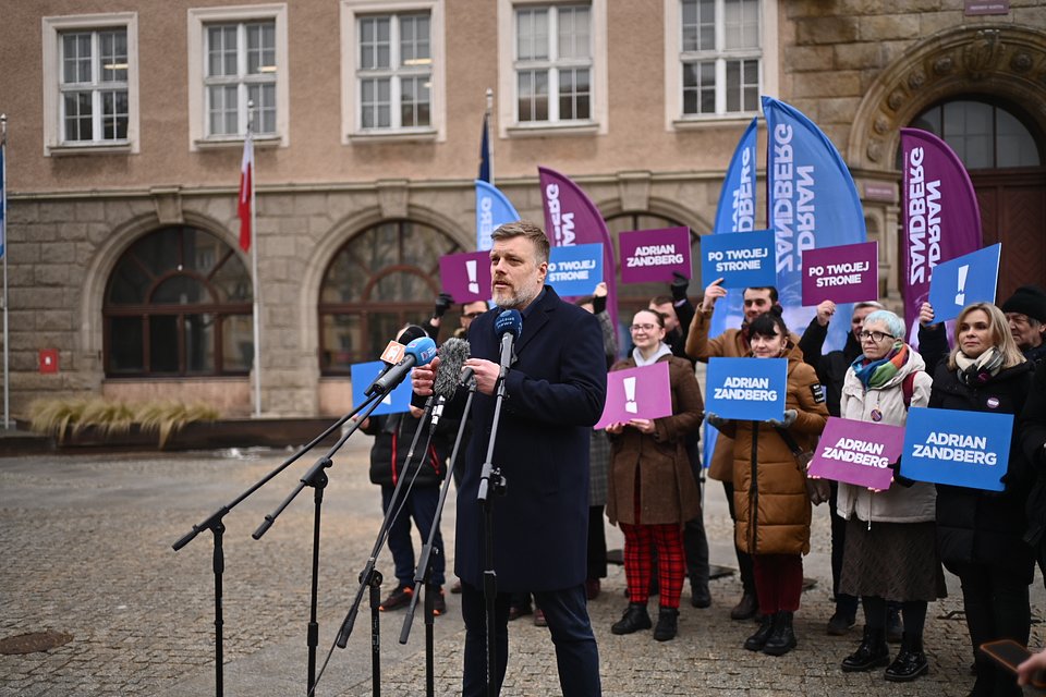 To kolejne ujęcie z konferencji Adriana Zandberga w Olsztynie, które doskonale ukazuje atmosferę wydarzenia – pełną energii, wsparcia i zaangażowania społecznego.  W tle widzimy działaczy i sympatyków Partii Razem, trzymających kolorowe tablice z hasłami kampanii: „Po twojej stronie”, „Adrian Zandberg” i symbolem wykrzyknika – znakiem alarmu, ale też determinacji. Charakterystyczne flagi w barwach fioletu i błękitu tworzą wyraziste tło podkreślające tożsamość polityczną wystąpienia.  Zandberg, stojący przy mikrofonach TVP Info, Polsat News i lokalnych stacji, przemawia z powagą i skupieniem. Budynek w tle – z flagą Polski i klasyczną architekturą – dodaje scenie formalnego charakteru i podkreśla wagę tematu.