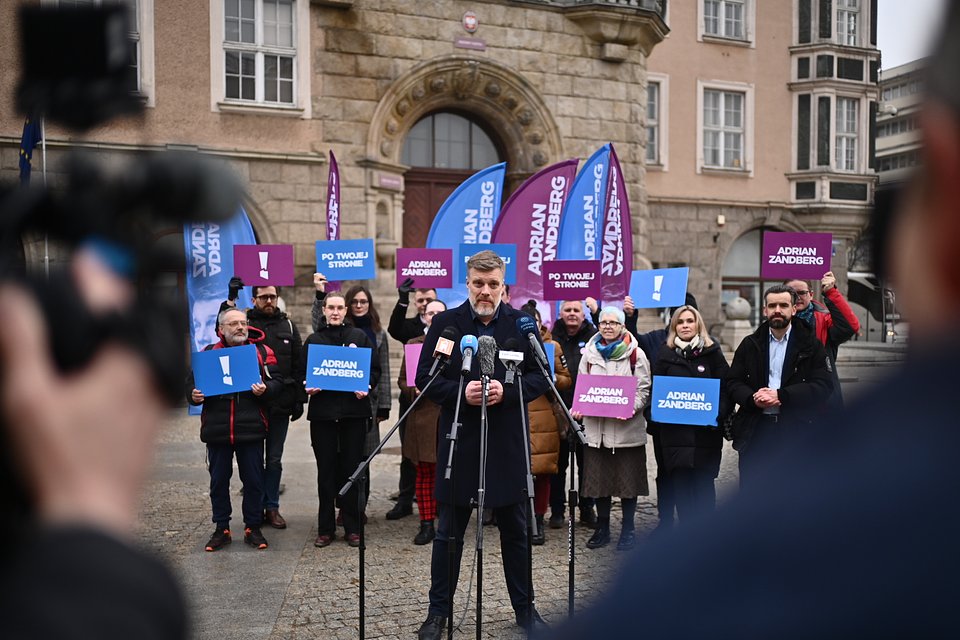 Na zdjęciu widzimy Adriana Zandberga podczas konferencji prasowej w Olsztynie, na tle zabytkowego gmachu (prawdopodobnie Ratusza). Zandberg przemawia do zgromadzonych dziennikarzy i mieszkańców, otoczony przez osoby trzymające banery z hasłami: „Adrian Zandberg”, „Po twojej stronie” oraz symbolem wykrzyknika – znakiem kampanii.  Flagi w barwach fioletu i błękitu, z imieniem i nazwiskiem lidera, wznoszą się za jego plecami, tworząc spójną oprawę wizualną wydarzenia. Wśród uczestników widać osoby dobrze znane z działalności w Partii Razem, w tym Bartosza Grucelę.  Zdjęcie ukazuje dynamiczny moment kampanii, w którym polityk lewicy zabiera głos w przestrzeni publicznej, podkreślając przekaz obecności „po stronie ludzi” – hasło szczególnie widoczne w trzymanych tablicach i emocjonalnym tonie wydarzenia.
