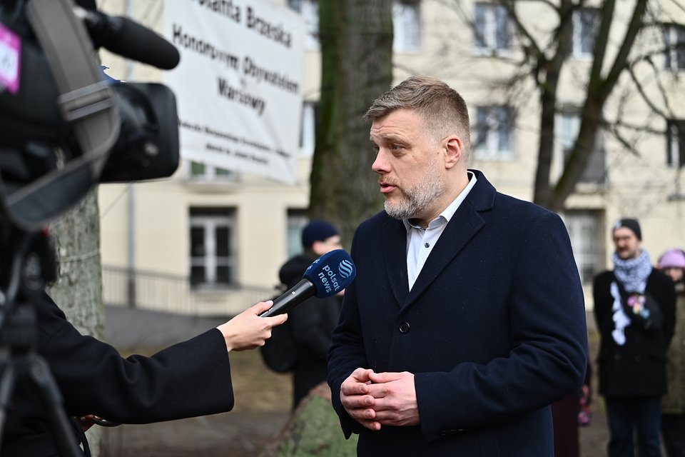 Na zdjęciu widzimy Adriana Zandberga udzielającego wywiadu dziennikarce Polsat News, tuż po uroczystościach upamiętniających 14. rocznicę śmierci Jolanty Brzeskiej. Tło stanowi baner z napisem „Jolanta Brzeska – Honorowa Obywatelka Warszawy”, co podkreśla symboliczny charakter miejsca.  Zandberg, ubrany elegancko w ciemny płaszcz, wygląda na skupionego i poruszonego, co koresponduje z powagą tematu. Jolanta Brzeska, działaczka ruchów lokatorskich, stała się symbolem walki o prawa mieszkańców – a jej tragiczna śmierć wciąż budzi silne emocje społeczne.  To zdjęcie oddaje atmosferę pamięci i zaangażowania politycznego w sprawy społeczne oraz walkę o sprawiedliwość lokatorską.