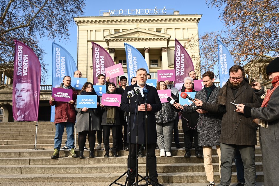 Na zdjęciu widzimy Adriana Zandberga przemawiającego podczas konferencji prasowej na schodach przed budynkiem Teatru Polskiego w Poznaniu, nad którym góruje słowo „WOLNOŚĆ” – znaczący i symboliczny kontekst dla wydarzenia politycznego.  Za Zandbergiem stoją osoby trzymające plansze z hasłami kampanii: „Adrian Zandberg”, „Po Twojej stronie” oraz znakiem „!”. Obok niego widoczni są dziennikarze z mikrofonami reprezentujący m.in. TVP Info, RMF FM i Polsat News. Tło stanowią charakterystyczne flagi Razem w kolorach fioletowym i niebieskim.  To zdjęcie ukazuje atmosferę zaangażowania i politycznego przekazu w przestrzeni publicznej, w centrum jednego z ważniejszych miast Polski.