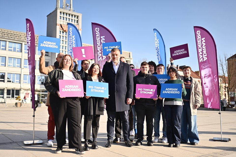 Na zdjęciu widzimy Adriana Zandberga pozującego do wspólnego zdjęcia z grupą sympatyków i sympatyczek w Koszalinie. To scena z kampanii wyborczej – uczestnicy trzymają w rękach plansze z hasłami: „Adrian Zandberg”, „Po Twojej stronie” oraz „!”. W tle stoją flagi wyborcze Razem z nazwiskiem kandydata.  Zdjęcie zostało wykonane na otwartej przestrzeni miejskiej, na tle nowoczesnych budynków i charakterystycznej wieży zegarowej. Kompozycja obrazu podkreśla zaangażowanie uczestników i jedność przekazu kampanijnego. Fotografia autorstwa Dawida Majewskiego z Razem.