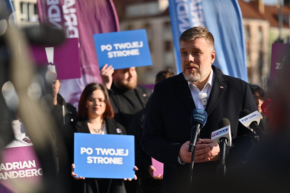 Na zdjęciu widzimy Adriana Zandberga podczas konferencji prasowej w Koszalinie. Przemawia on do mediów – przed nim ustawiono mikrofony m.in. stacji TVP Info, Polsat News, TV Max i innych. Za nim znajdują się członkowie jego zespołu oraz sympatycy trzymający charakterystyczne niebieskie i fioletowe tabliczki z hasłami kampanii: „Po Twojej stronie” oraz „Adrian Zandberg”.  W tle widoczne są flagi wyborcze i zabudowa miejska. Zandberg, ubrany w ciemny płaszcz, mówi z zaangażowaniem – cała scena ma dynamiczny, ale uporządkowany charakter typowy dla kampanii wyborczych.  Zdjęcie uchwyciło moment mobilizacji lokalnych sympatyków i przekazu politycznego skierowanego do mieszkańców Koszalina.