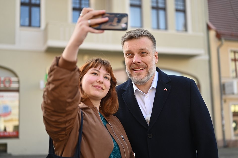 Na zdjęciu widzimy Adriana Zandberga w Zielonej Górze, pozującego do wspólnego selfie z młodą kobietą. Oboje uśmiechają się, a kobieta trzyma w górze telefon, robiąc zdjęcie. W tle widać jasną elewację miejskiego budynku z dużymi oknami i zaokrąglonymi łukami przy wejściach — typową dla zabytkowego centrum miasta. Atmosfera na zdjęciu jest przyjazna i swobodna, a Zandberg prezentuje się w granatowym płaszczu z przypiętą biało-czerwoną flagą.  To ujęcie ukazuje bezpośredni kontakt polityka z mieszkańcami, podkreślając jego dostępność i pozytywne nastawienie podczas kampanii wyborczej.