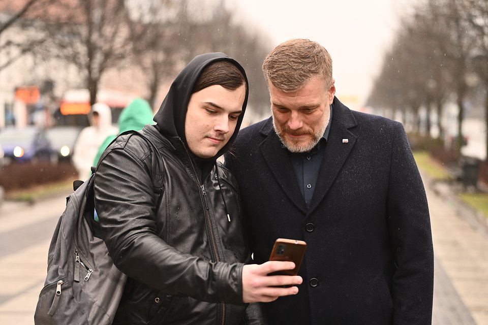 Na zdjęciu widzimy Adriana Zandberga rozmawiającego z młodym mężczyzną podczas spaceru ulicą w Częstochowie. Obaj skupieni są na ekranie telefonu – wygląda to na moment wspólnego przeglądania zdjęcia lub przygotowywania selfie. Zandberg ubrany jest w granatowy płaszcz, jego rozmówca – w czarną skórzaną kurtkę z kapturem i plecakiem.  Tło stanowi alejka miejska z rzędami drzew i rozmytymi sylwetkami przechodniów w płaszczach i kapturach – sugerującymi chłodny, deszczowy dzień. Zdjęcie oddaje bezpośredni, codzienny kontakt polityka z mieszkańcami i ich zainteresowanie jego obecnością.