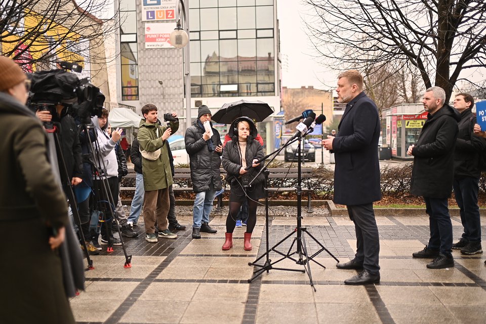 Na zdjęciu widzimy Adriana Zandberga przemawiającego podczas konferencji prasowej na ulicy w Częstochowie. Stoi przed zestawem mikrofonów, a naprzeciw niego zgromadzili się dziennikarze i kamerzyści. Wśród nich widoczna jest kobieta trzymająca mikrofon i parasol, osoby z aparatami oraz kilka kamer telewizyjnych.  W tle znajdują się miejskie budynki, witryny sklepowe i charakterystyczny szary gmach z napisem „LUZ”. Na dalszym planie widać też czerwono-żółty kiosk. Po prawej stronie kadru stoją członkowie zespołu Zandberga, w tym Maciej Konieczny – trzymający tabliczkę z napisem „Po Twojej stronie”.  Zdjęcie ukazuje polityczną codzienność i medialne zainteresowanie działaniami lidera partii Razem.