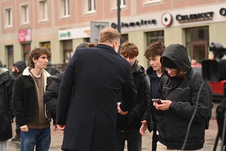 Na zdjęciu widzimy Adriana Zandberga w trakcie rozmowy z grupą młodych wyborców na ulicy w Mielcu. Polityk ubrany w ciemny płaszcz pochyla się i podaje dłoń jednej z osób, nawiązując bezpośredni kontakt. Scena odbywa się w miejskiej przestrzeni – w tle widoczne są sklepy i punkty usługowe, w tym m.in. optyk i jubiler, co sugeruje, że zdjęcie zostało wykonane w centrum miasta.  Młodzi ludzie stoją wokół Zandberga – część z nich słucha uważnie, jedna osoba korzysta z telefonu, być może robiąc zdjęcie lub nagrywając film. W kadrze widać też fragment kamery telewizyjnej, co sugeruje, że spotkaniu towarzyszyła obecność mediów.  Zdjęcie oddaje atmosferę zaangażowania i autentycznego zainteresowania – to scena polityki bliskiej ludziom, w której kandydat nie przemawia z mównicy, lecz rozmawia wprost z obywatelami, twarzą w twarz.