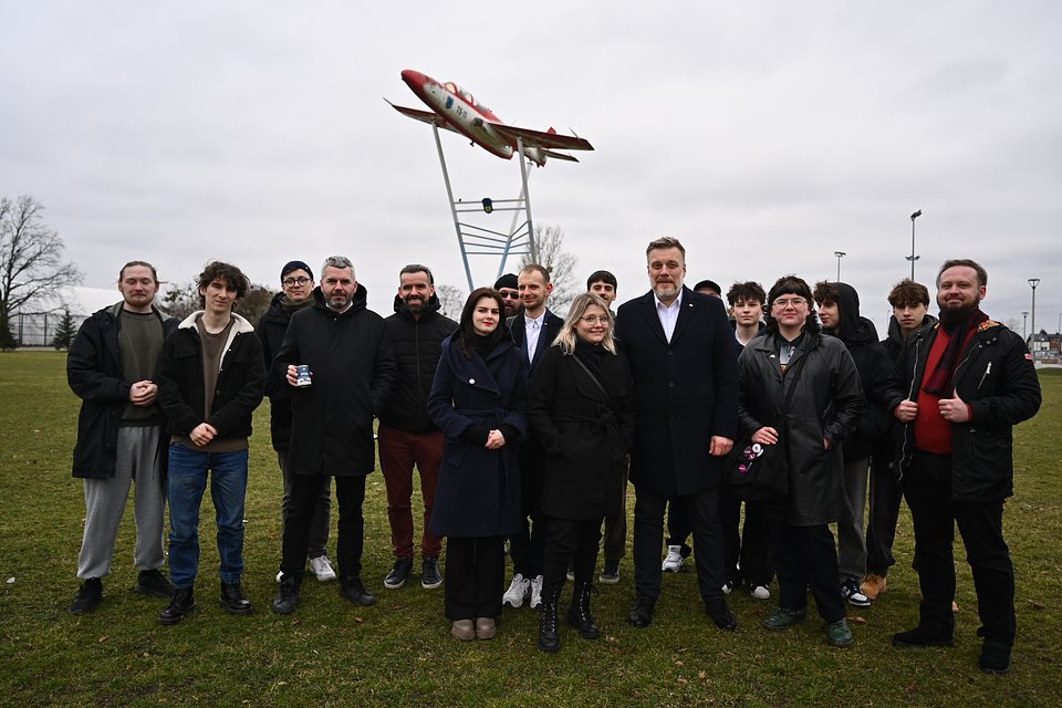 Na zdjęciu widzimy Adriana Zandberga stojącego w centrum grupy młodych ludzi i działaczy lokalnych, podczas spotkania z wyborcami w Mielcu. Tłem tej plenerowej sceny jest charakterystyczna konstrukcja z samolotem odrzutowym – pomnik-symbol nawiązujący do przemysłowego dziedzictwa miasta i lotniczej historii Mielca.  Zandberg, ubrany w czarny płaszcz i białą koszulę, stoi w pierwszym rzędzie, uśmiechnięty, w otoczeniu m.in. Macieja Koniecznego oraz Aleksandry Owcy – współprzewodniczącej Razem. Resztę grupy stanowią sympatycy i sympatyczki w różnym wieku – część z nich to osoby młode, ubrani nieformalnie, co podkreśla swobodny, bezpośredni charakter tego spotkania.  Zdjęcie emanuje energią wspólnoty i zaangażowania, a obecność symbolicznego samolotu w tle nadaje mu lokalny, tożsamościowy kontekst. To moment spotkania z ludźmi, rozmowy o sprawach regionu i wspólnej politycznej nadziei.