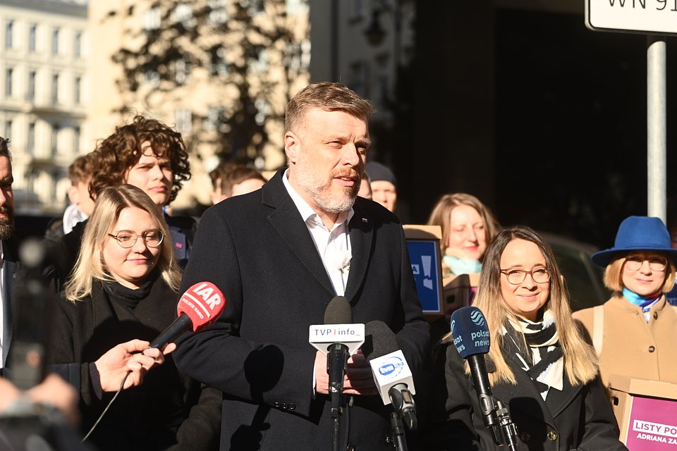 Na zdjęciu widzimy Adriana Zandberga podczas konferencji prasowej zorganizowanej przed siedzibą Państwowej Komisji Wyborczej (PKW), tuż przed formalną rejestracją jego kandydatury. Zandberg stoi na czele grupy współpracowników i współpracowniczek, w tym posłanek partii Razem – m.in. Marceliny Zawiszy i Pauliny Matysiak – oraz działaczy młodszego pokolenia.  Ubrany w ciemny płaszcz i białą koszulę, mówi do zgromadzonych dziennikarzy, przy kilku mikrofonach należących do różnych redakcji: TVP Info, Polsat News, IAR, TVN24. W tle widać transparenty z hasłami kampanii, m.in. „Listy poparcia Adriana Zandberga”, a także sympatyków i sympatyczki jego komitetu.  Zdjęcie ukazuje moment oficjalny, ale jednocześnie radosny i pełen energii – słońce pada mocno na twarze uczestników, co nadaje scenie blasku i atmosfery nadziei. To symboliczny krok w kampanii, pokazujący jedność zespołu i gotowość do walki o najwyższy urząd w państwie.