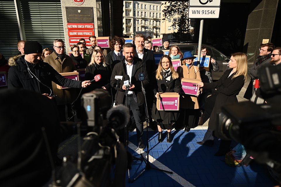 Na zdjęciu widzimy Bartosza Grucelę, szefa sztabu wyborczego Adriana Zandberga, przemawiającego podczas konferencji prasowej przed siedzibą Państwowej Komisji Wyborczej (PKW) w Warszawie. Stoi on przy mikrofonach należących do ogólnopolskich mediów – m.in. TVP Info, Polsat News, TVN24 i IAR – kierując swoje słowa do dziennikarzy i opinii publicznej.  Za nim, w zwartym szeregu, stoją członkinie i członkowie komitetu wyborczego kandydata na prezydenta Adriana Zandberga, w tym Adrian Zandberg, Marcelina Zawisza i Paulina Matysiak. Wiele osób trzyma pudełka i plansze z hasłami kampanii: „Listy poparcia Adriana Zandberga”, co symbolizuje złożenie wymaganej liczby podpisów poparcia. Na czerwonej tablicy budynku widoczne są oficjalne oznaczenia PKW i Krajowego Biura Wyborczego.  Zdjęcie ukazuje kluczowy moment w kampanii – ogłoszenie formalnego zgłoszenia kandydatury, przy pełnym zaangażowaniu zespołu. Scena jest dynamiczna i profesjonalna, oddaje powagę chwili, ale też energię i mobilizację całego środowiska.