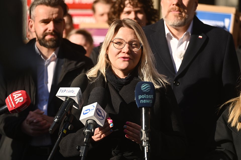 Na zdjęciu widzimy Aleksandrę Owcę, współprzewodniczącą partii Razem, przemawiającą podczas konferencji prasowej zorganizowanej przed siedzibą Państwowej Komisji Wyborczej (PKW). Owca stoi w centrum kadru, uśmiechnięta i pewna siebie, mówiąc do zgromadzonych przedstawicieli mediów – widać mikrofony należące do największych stacji informacyjnych: Polsat News, TVP Info, TVN24, IAR.  W tle, nieco rozmyci, stoją inni politycy partii Razem, w tym Bartosz Grucela i Adrian Zandberg, co podkreśla wspólnotowy charakter wystąpienia. Słońce mocno oświetla twarze uczestników, co nadaje scenie energii i optymizmu.  Zdjęcie ukazuje Aleksandrę Owcę jako jedną z twarzy kampanii, reprezentującą nową generację polityczek Razem – zdecydowaną, komunikatywną i zorientowaną na przekaz. To moment zbiorowej mobilizacji i siły zespołowej, który towarzyszy oficjalnemu zgłoszeniu kandydatury Adriana Zandberga.