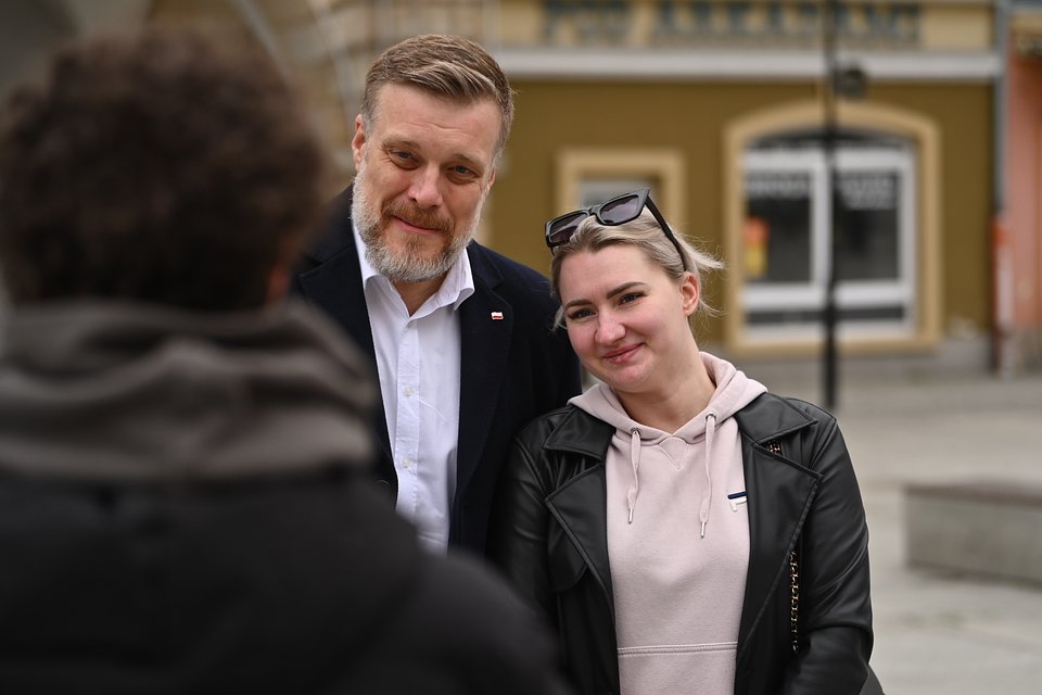 Na zdjęciu widzimy Adriana Zandberga podczas spotkania z mieszkańcami Nysy. Polityk stoi obok młodej kobiety, która uśmiecha się do osoby wykonującej zdjęcie – to zapewne moment uchwycony po krótkiej rozmowie lub prośbie o wspólne zdjęcie. Zandberg również delikatnie się uśmiecha, tworząc serdeczną i przyjazną atmosferę.  Kobieta ubrana jest w jasnoróżową bluzę z kapturem i czarną ramoneskę, na głowie ma okulary przeciwsłoneczne. Zandberg nosi ciemny płaszcz i białą koszulę, a przy klapie jego płaszcza widoczna jest przypinka z polską flagą. W tle znajdują się klasycystyczne kamienice w stonowanych kolorach, co sugeruje rynek lub centralny plac Nysy.  To zdjęcie ukazuje bardziej osobisty, ludzki wymiar kampanii – bez mikrofonów i tłumu, za to z bezpośrednim kontaktem i ciepłym gestem obecności.
