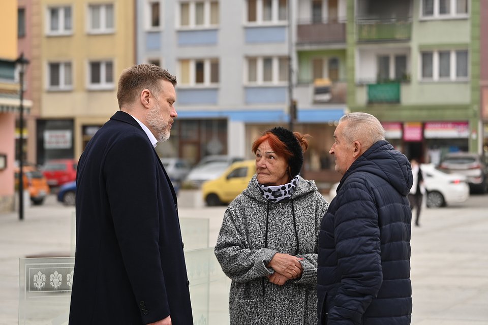 Na zdjęciu widzimy Adriana Zandberga w trakcie rozmowy z dwojgiem starszych mieszkańców Nysy. Scena rozgrywa się na miejskim rynku lub placu – w tle widoczne są kolorowe elewacje bloków z lokalami usługowymi na parterze, zaparkowane samochody i przechodnie.  Zandberg, ubrany w ciemny płaszcz i białą koszulę, z powagą i skupieniem słucha rozmówców – kobiety w szarej bluzie z czarno-białym kominem i czarną opaską na włosach oraz mężczyzny w granatowej kurtce pikowanej. Ich postawa sugeruje zaangażowanie i otwartość na rozmowę o lokalnych sprawach.  Zdjęcie ukazuje spokojną, codzienną scenę spotkania polityka z mieszkańcami – bez zbędnego patosu, za to z naciskiem na uważne słuchanie i bezpośredni kontakt twarzą w twarz. To obraz bliskiej ludzi kampanii, rozgrywającej się w realnym krajobrazie małego miasta.