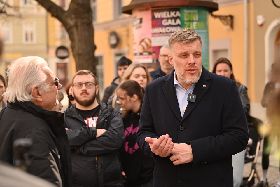 Na zdjęciu widzimy Adriana Zandberga podczas spotkania z mieszkańcami Świdnicy. Zandberg, ubrany w granatowy płaszcz i jasnoniebieską koszulę, rozmawia z grupą osób zgromadzonych na ulicy. Przy klapie płaszcza widoczna jest przypinka z flagą Polski oraz mikrofon przypięty do kołnierza, co sugeruje, że rozmowy są rejestrowane lub transmitowane.  W centrum kadru Zandberg zwraca się do starszego mężczyzny z siwymi włosami i okularami przeciwsłonecznymi. W tle stoją słuchacze – osoby w różnym wieku, uważnie przysłuchujące się rozmowie. Za grupą widać staromiejską architekturę Świdnicy: kolorowe kamienice, słup ogłoszeniowy z plakatami (m.in. „Dni Ziemi”, „Wielka Gala”), a także dekoracyjne detale fasad.  Atmosfera jest otwarta i pełna bezpośredniego kontaktu – to scena z politycznego spaceru lub otwartego spotkania w przestrzeni publicznej, ukazująca bliskość polityka z ludźmi oraz gotowość do rozmowy na ich warunkach.