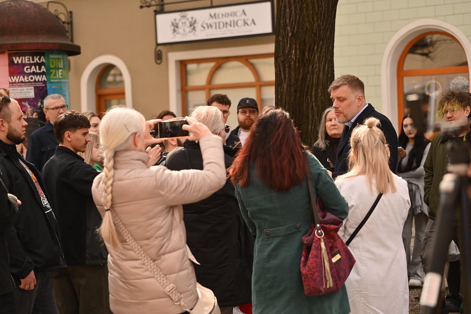 Na zdjęciu widzimy Adriana Zandberga w trakcie spotkania z mieszkańcami, które odbywa się na tle miejskiej architektury – najpewniej w Świdnicy, co potwierdza widoczny szyld „Mennica Świdnicka”. Zandberg stoi po prawej stronie kadru, ubrany w granatowy płaszcz i jasną koszulę, rozmawia z osobami zebranymi wokół niego. Jego postawa i gesty wskazują na skupienie i powagę, jakby odpowiadał na ważne pytanie.  Wokół zgromadzili się mieszkańcy – osoby w różnym wieku i stylu. Jedna z kobiet w jasnym płaszczu fotografuje lub nagrywa spotkanie telefonem. W tłumie wyróżniają się osoby z białymi lub zielonymi włosami, czarnymi ubraniami i wyrazistym makijażem, co dodaje scenie autentyczności i różnorodności. W tle widoczne są miejskie szyldy i słup ogłoszeniowy z plakatami wydarzeń lokalnych.  Zdjęcie przedstawia Zandberga w bezpośrednim kontakcie z wyborcami – to scena z ulicznej kampanii, pokazująca bliskość polityka wobec ludzi i realnych rozmów, bez bariery sceny czy mikrofonu.