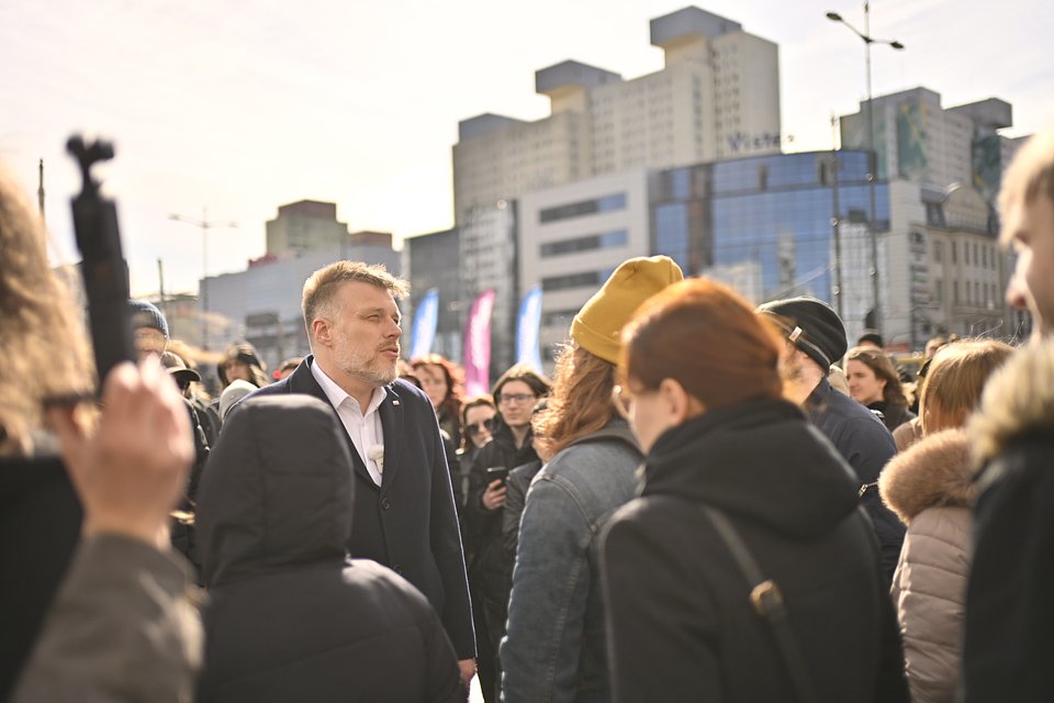 Na zdjęciu widzimy Adriana Zandberga podczas spotkania z mieszkańcami Łodzi, w otwartej przestrzeni miejskiej. Polityk stoi w centrum grupy osób, ubrany w ciemny płaszcz i białą koszulę – słucha uważnie lub odpowiada na pytania zebranych wokół niego ludzi.  W tle widać charakterystyczną nowoczesną zabudowę centrum Łodzi, z rozpoznawalnymi biurowcami i galerią handlową, na której fasadzie widnieje szyld „West…”. Na dalszym planie widać także flagi kampanijne w kolorach niebieskim i fioletowym z napisem „Adrian Zandberg”, sugerujące, że wydarzenie jest częścią jego trasy wyborczej.  Atmosfera zdjęcia jest dynamiczna i skupiona, pełna bezpośrednich rozmów – to scena, która podkreśla zaangażowanie w dialog obywatelski i obecność w przestrzeni miejskiej, wśród codziennych przechodniów i wyborców.