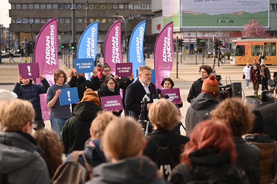 Na zdjęciu widzimy Adriana Zandberga przemawiającego podczas konferencji prasowej w Łodzi, zorganizowanej na otwartej przestrzeni miejskiej – najprawdopodobniej w rejonie skrzyżowania Marszałków (tzw. „styk Piotrkowskiej i Mickiewicza”), rozpoznawalnym punkcie miasta.  Zandberg stoi przy mikrofonach mediów, zwrócony do zebranej publiczności. Ubrany jest w ciemny płaszcz i białą koszulę, mówi z zaangażowaniem. Za nim stoją działacze i działaczki kampanii z tablicami w kolorach fioletowym i niebieskim, z hasłami: „Adrian Zandberg”, „Po twojej stronie” oraz symbolem wykrzyknika – charakterystycznym dla partii Razem. W tle widoczne są nowoczesne i modernistyczne budynki, tramwaj oraz miejska przestrzeń reklamowa, która wzmacnia kontekst wielkomiejski wydarzenia.  Przed Zandbergiem zebrała się spora grupa słuchaczy – młodych ludzi i dziennikarzy, z kamerami i aparatami. Atmosfera jest energiczna, zaangażowana i dobrze zorganizowana – widać, że to część oficjalnej kampanii wyborczej prowadzonej z myślą o otwartości, dialogu i obecności w centrum życia społecznego.