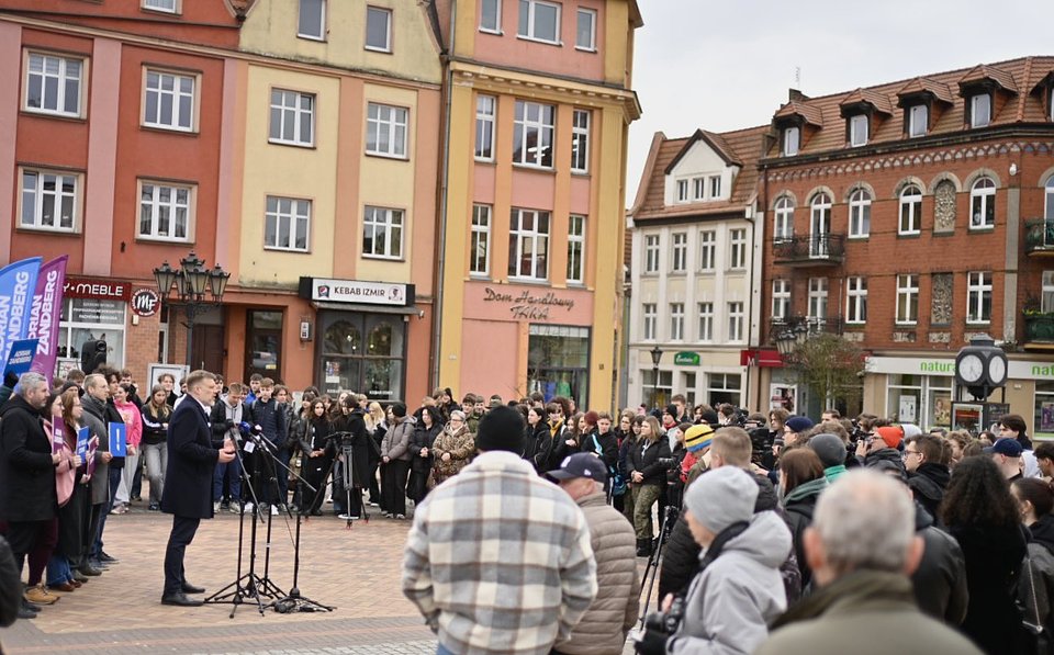 Na zdjęciu widzimy Adriana Zandberga przemawiającego podczas spotkania z wyborcami na rynku w Chojnicach. Stoi przy zestawie mikrofonów, otoczony przez członków swojego sztabu i sympatyków, którzy trzymają tablice kampanijne w kolorach fioletowym i niebieskim z hasłami „Adrian Zandberg” i symbolem wykrzyknika.  Przed nim zgromadził się spory tłum słuchaczy – mieszkańców Chojnic – uważnie słuchających przemówienia. Uczestnicy spotkania są ubrani zimowo, co sugeruje chłodniejszy dzień. W tle widoczna jest charakterystyczna zabudowa rynku – kolorowe kamienice, lokale usługowe oraz miejskie zegary.  Atmosfera zdjęcia jest żywa i obywatelska – to typowa scena z kampanii prowadzonej w przestrzeni publicznej, nastawionej na bezpośredni kontakt z ludźmi, wsłuchiwanie się w ich głosy i prezentowanie programowych propozycji w sercu lokalnej społeczności.