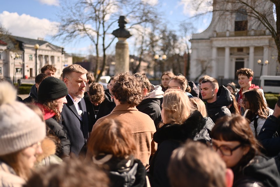 Na zdjęciu widzimy Adriana Zandberga podczas spotkania z mieszkańcami w przestrzeni miejskiej – na placu w Siedlcach. Otoczony jest przez tłum młodych ludzi, z którymi rozmawia w bezpośredni, zaangażowany sposób. Zandberg ma na sobie ciemny płaszcz i jasną koszulę, wygląda na skupionego i zainteresowanego rozmową.  W tle widoczna jest klasycystyczna architektura – biały budynek z kolumnowym portykiem oraz pomnik z popiersiem na cokole, co wskazuje na centralną część miasta. Pogoda jest słoneczna, niebo jasne, a ludzie ubrani są w zimowe i przejściowe kurtki, co sugeruje chłodniejszy, wiosenny dzień.  Zdjęcie oddaje atmosferę otwartości i dialogu – polityk rozmawiający twarzą w twarz z obywatelami, bez dystansu, w otwartym kręgu rozmówców, co wzmacnia przekaz o bliskości i dostępności w kampanii wyborczej.